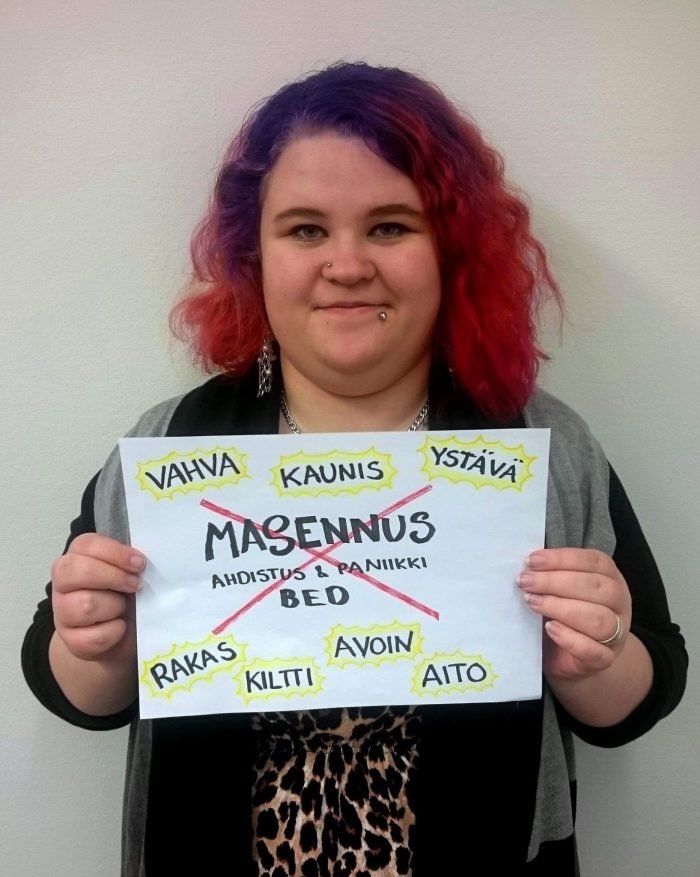 Krista pitelee kylttiä, jossa sana masennus on viivattu yli ja korvattu positiivisilla sanoilla kuten "rakas", "ystävä" ja "aito".
